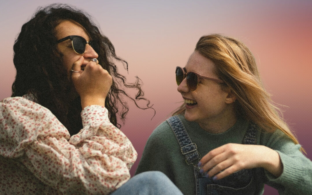 Photo of two young women laughing together outdoors in the sunshine. Both wear sunglasses. Image provided by BABI at Unsplash