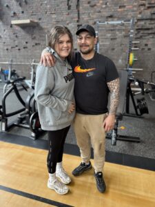 male and female partners stand next to each other in the gym smiling at camera