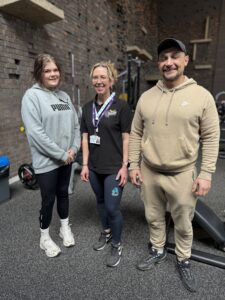male and female physical activity clients smile stood next to fgs advisor in the gym